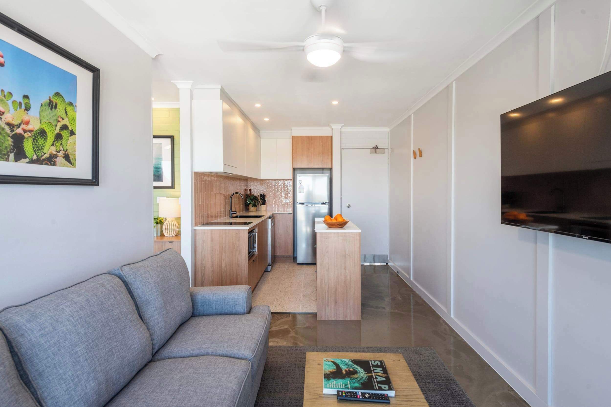 A modern living room with a grey sofa, a coffee table with a book, a kitchen area with a fridge, and a TV mounted on the wall.