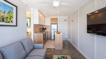 A modern living room with a grey sofa, a coffee table with a book, a kitchen area with a fridge, and a TV mounted on the wall.