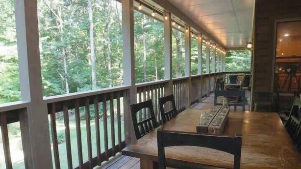 A covered outdoor dining area with wooden tables and chairs, large windows, and a view of a forested area.