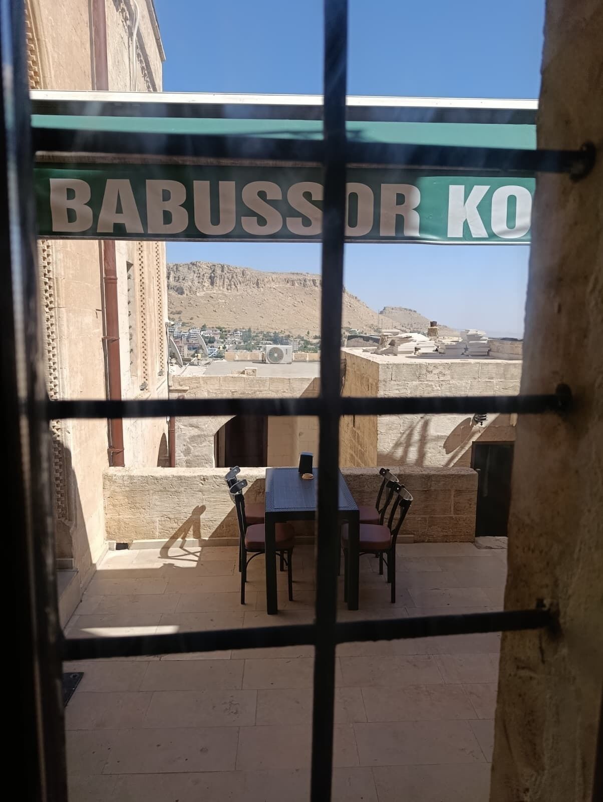A view through a window with a sign reading "BABUSSOR KO" and a table with chairs outside.