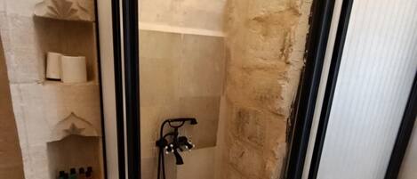 Ein kleiner, abgeschlossener Duschbereich mit Fliesenboden und Duschkopf. Regale mit Toilettenartikeln und ein Besen stehen auf dem Boden.