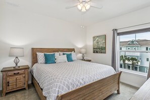 A bedroom with a wooden bed, two bedside tables, a ceiling fan, a window with a view of buildings, and a painting on the wall. - Private Beach Club| Two pools| Free Rounds of Golf| Free Community Shuttle| Tennis/Pickleball (Pensacola)