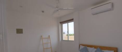 A bedroom with a bed, a ceiling fan, an air conditioning unit, a window, and a wooden ladder.