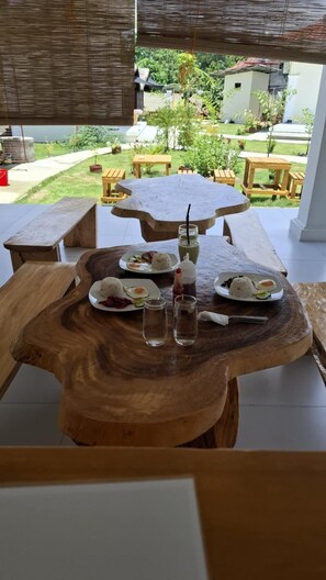 A wooden dining table with a unique design, surrounded by outdoor seating and a view of a garden and buildings in the background. - The Kubo Gardens (San Juan)