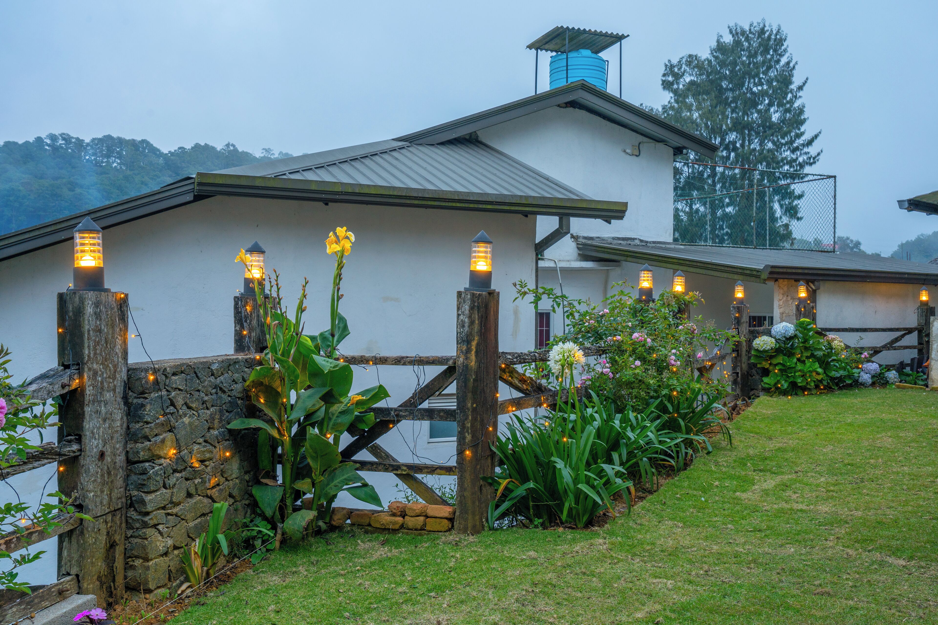 A rustic house with a stone wall and wooden fence, surrounded by lush greenery and flowering plants.