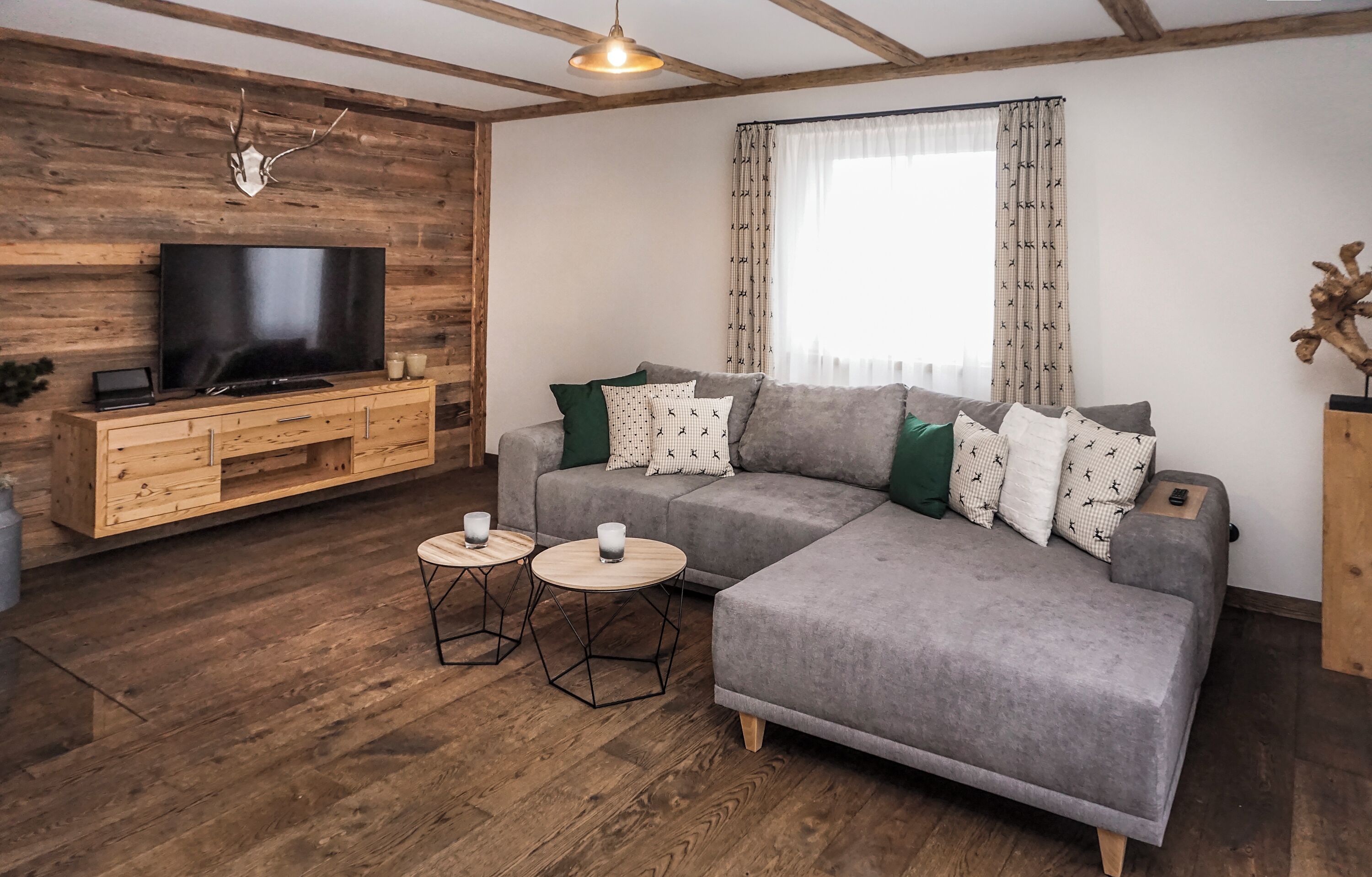 Una sala de estar moderna con un sofá gris, suelo de madera, un televisor sobre un mueble de madera y una ventana con cortinas estampadas.