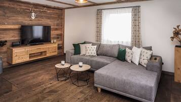 Una sala de estar moderna con un sofá gris, suelo de madera, un televisor sobre un mueble de madera y una ventana con cortinas estampadas.