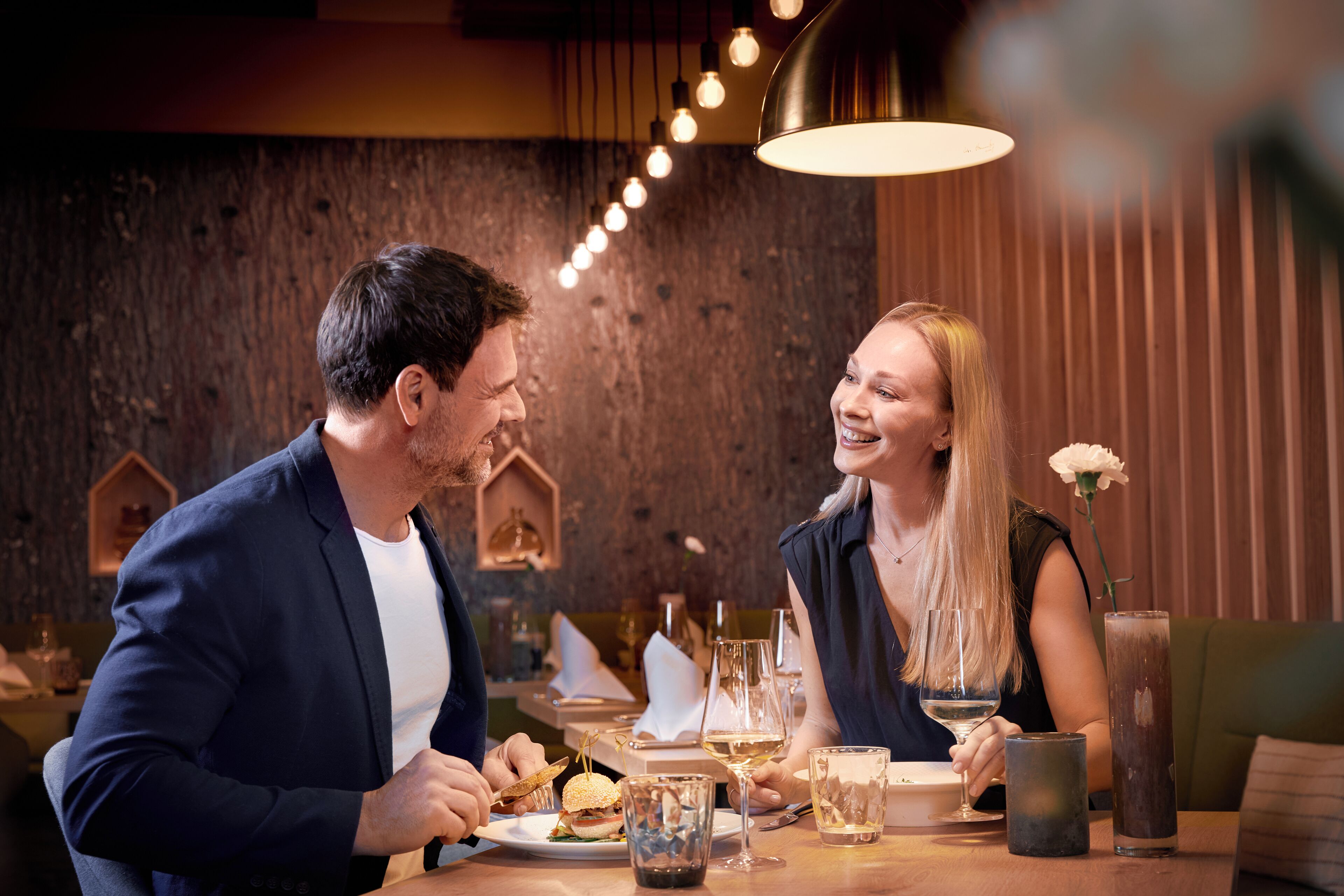 Un couple est assis à une table dans un restaurant dont le mur est fait de bois brut, décoré de guirlandes lumineuses, et sur laquelle est posé un vase contenant une fleur blanche.