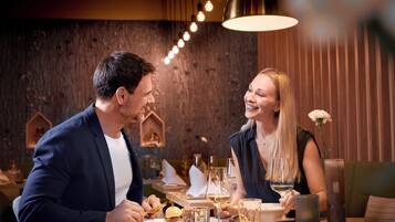 Un couple est assis à une table dans un restaurant dont le mur est fait de bois brut, décoré de guirlandes lumineuses, et sur laquelle est posé un vase contenant une fleur blanche.