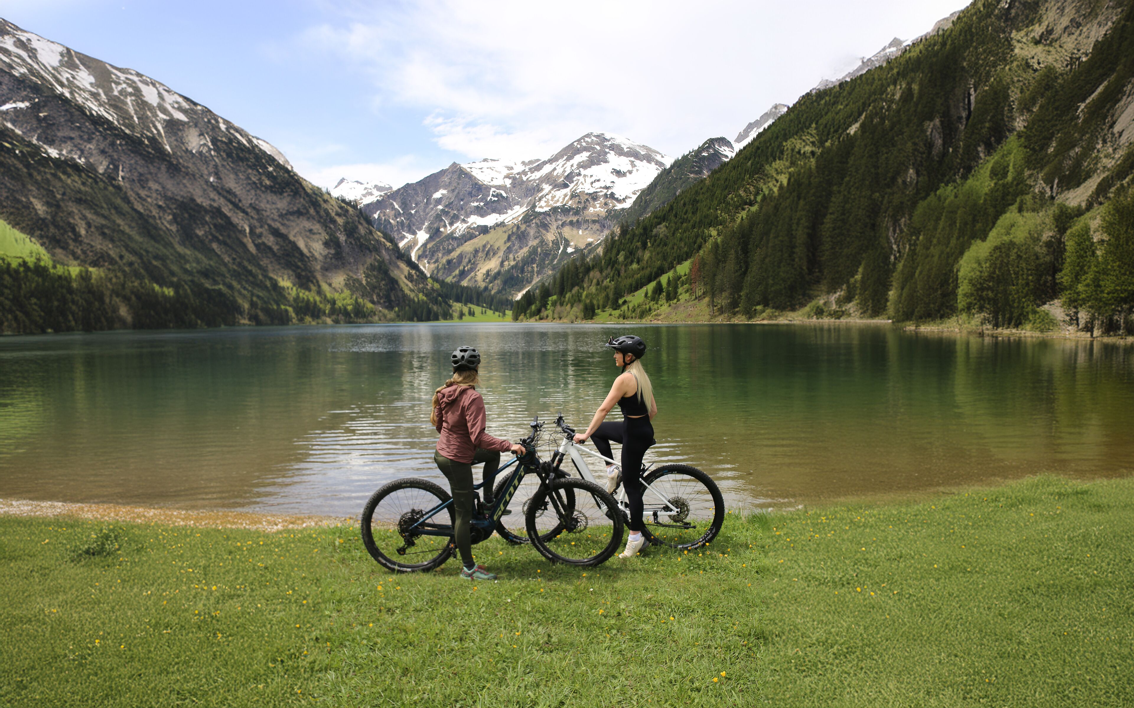 Two people on bicycles by a lake with mountains in the background.