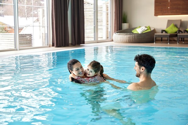An indoor swimming pool with clear blue water, large windows, and a comfortable seating area with cushions. - Alpin Lodges Oberjoch (Bad Hindelang)