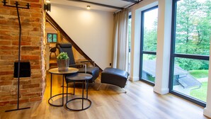 A modern interior with a brick wall, a wooden floor, a black leather sofa, a round table with a plant, and large windows offering a view of greenery. - Apartment 'Michels Mühle  - Pitt Sin Atelier' with Private Terrace, Private Garden and Wi-Fi (Brilon)