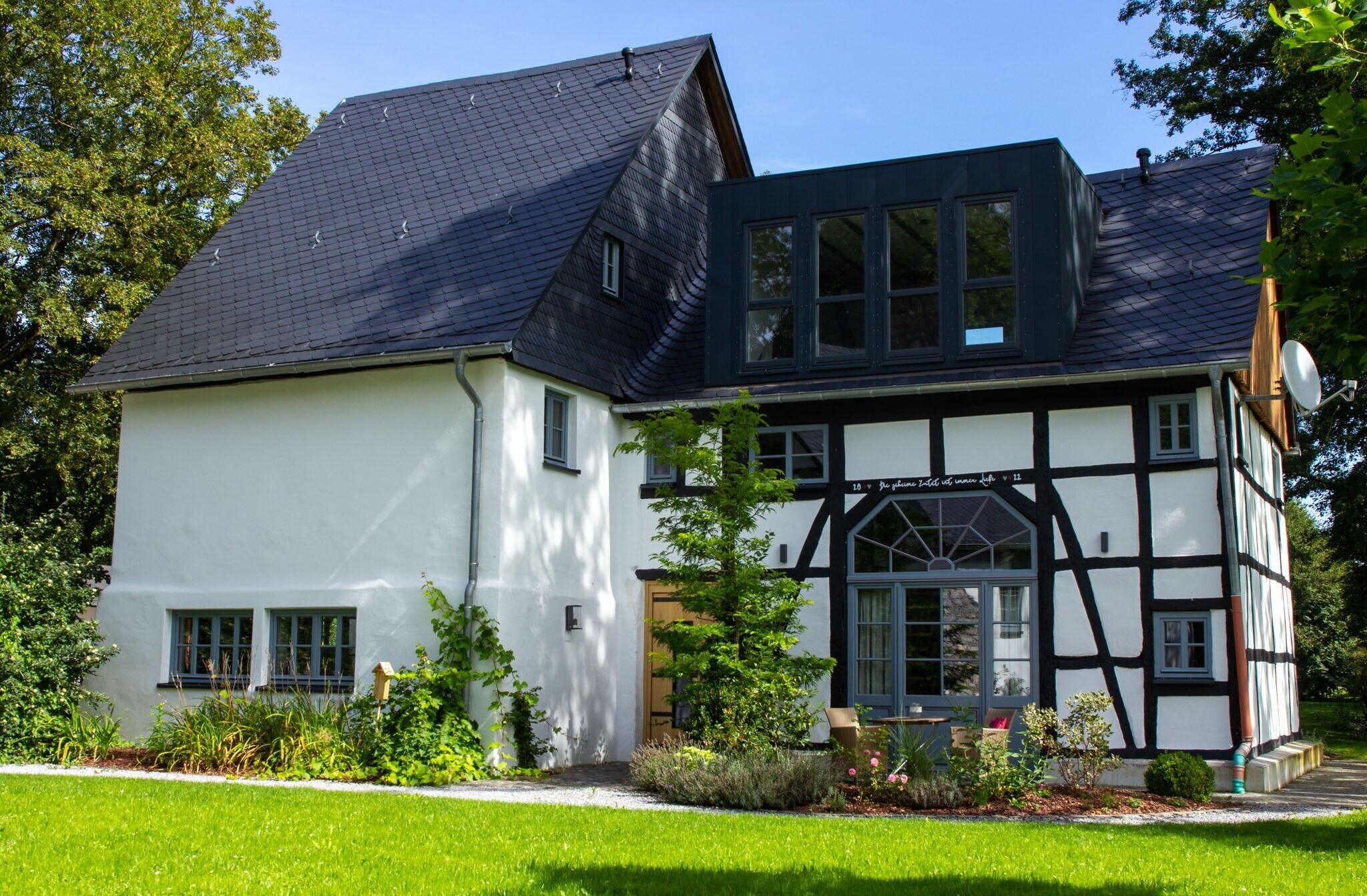 Uma casa de dois andares com fachada branca e telhado escuro, cercada por vegetação.