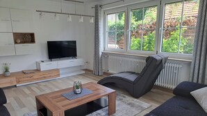 A modern living room with a flat-screen TV, a sofa, a coffee table, and a window with a view of greenery. - Apartment in the Ries with Wi-Fi (Wallerstein)