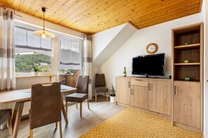 A room with a dining table, chairs, a TV, wooden cabinets, a clock, and a window with a view of greenery. - Apartment 'Ferienwohnung Typ 1' with Mountain View, Shared Terrace and Wi-Fi (Willingen (Upland))