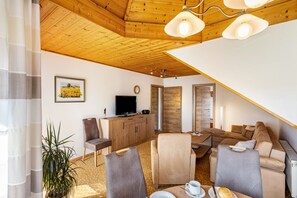 A modern living room with a wooden ceiling, a television, a dining area with a table and chairs, and a large window with curtains.