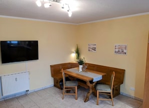 Ein Essbereich mit Holztisch, zwei Stühlen, einer Bank, einem an der Wand befestigten Fernseher und einer Fußbodenheizung.