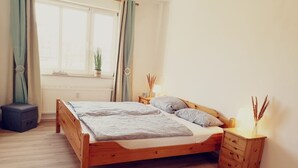 Quarto com cama de madeira, mesa de cabeceira com abajur, um pequeno banquinho e janela com cortinas.