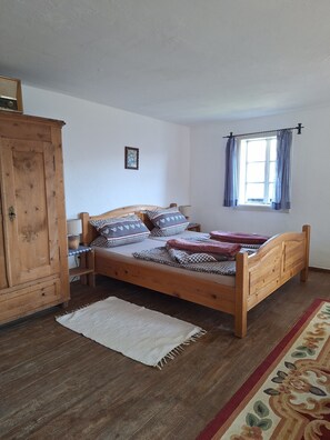 A wooden bed with patterned bedding, a wooden wardrobe, a small bedside table, a nightstand, a window with curtains, and a patterned rug on a wooden floor.