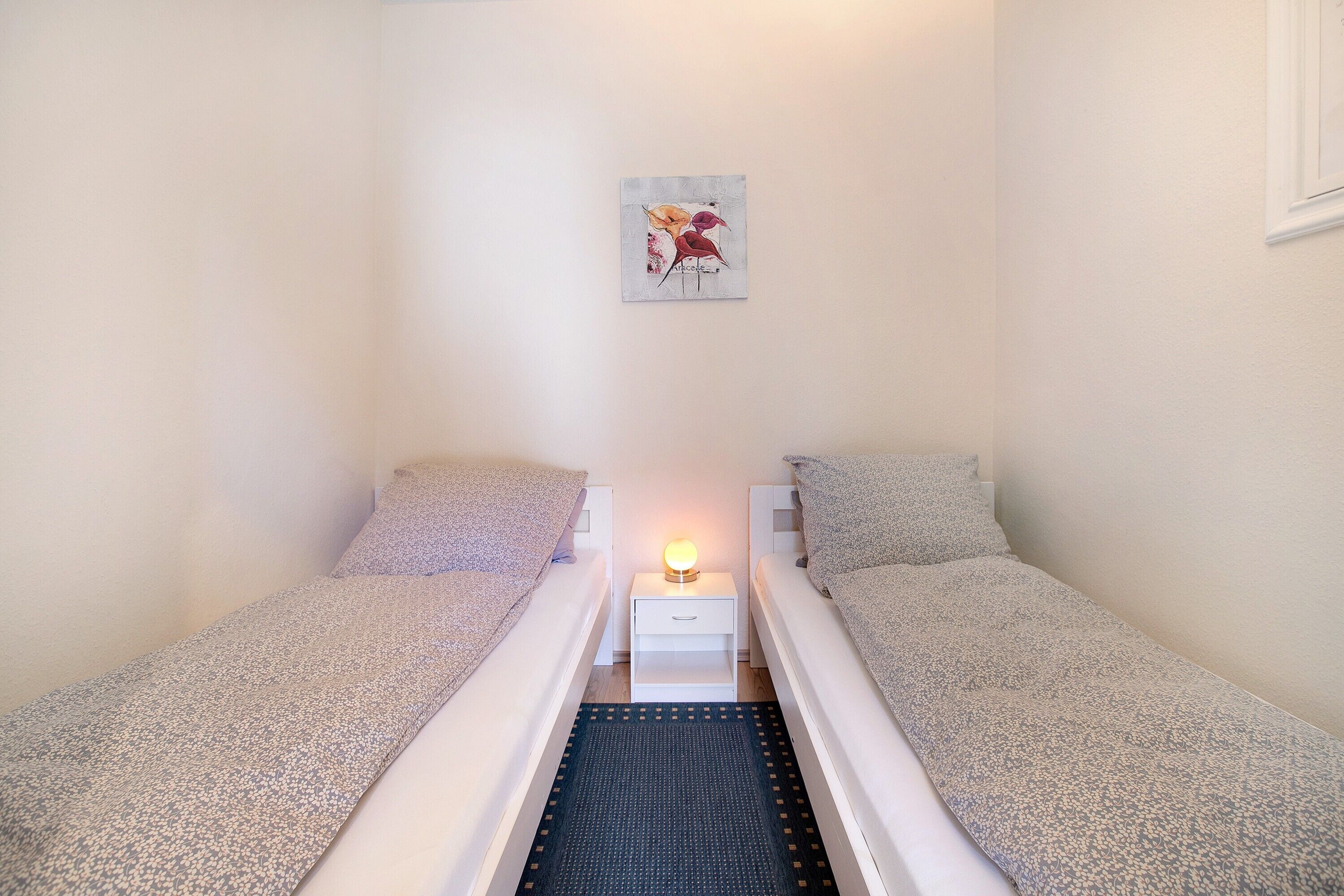 Een kleine, schone kamer met twee eenpersoonsbedden, een nachtkastje met een lamp en een kunstwerk aan de muur.