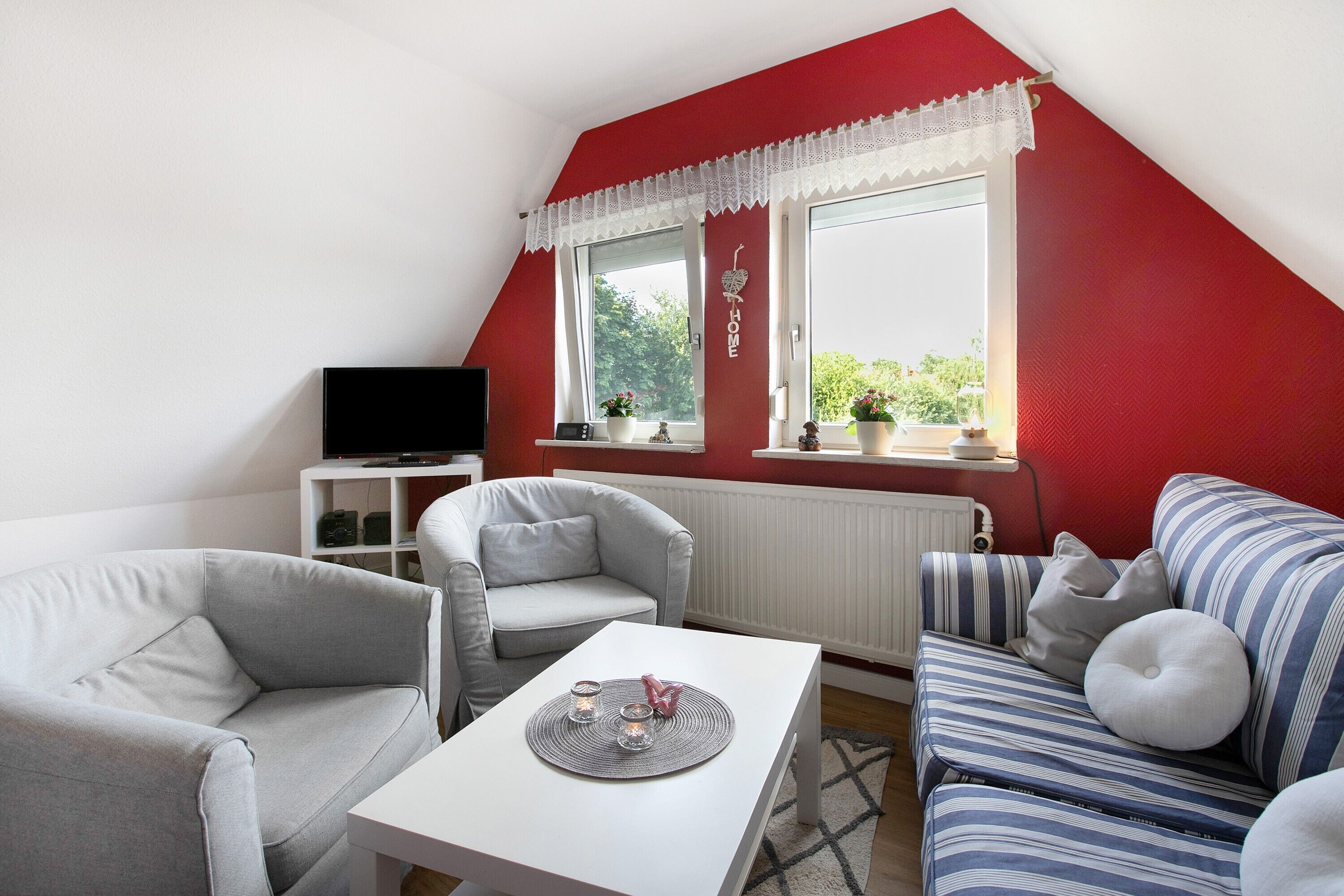 Una habitación con una pared roja, un radiador blanco, un sofá gris, un sofá a rayas, una mesa blanca y un televisor sobre un soporte.