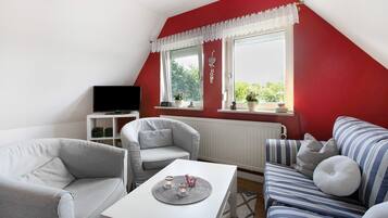Una habitación con una pared roja, un radiador blanco, un sofá gris, un sofá a rayas, una mesa blanca y un televisor sobre un soporte.