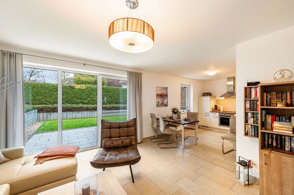 A modern living room with a sofa, a dining table with chairs, a kitchen area, and a bookshelf.