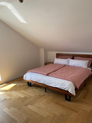 A bedroom with a wooden bed, white and pink bedding, wooden flooring, and a sloped ceiling.