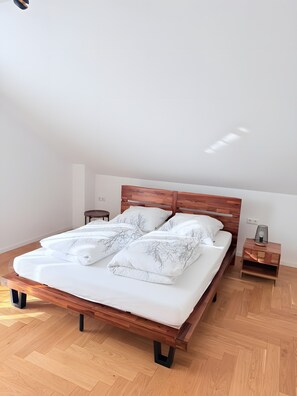 Quarto com estrutura de cama de madeira, roupa de cama branca, mesa de cabeceira com abajur e piso de madeira em padrão herringbone.