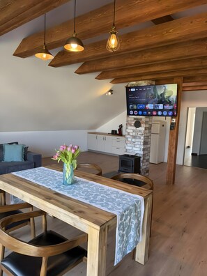 Uma sala de estar moderna com teto de madeira, uma mesa de jantar com cadeiras, uma TV fixada numa parede de tijolos e um sofá acolhedor.