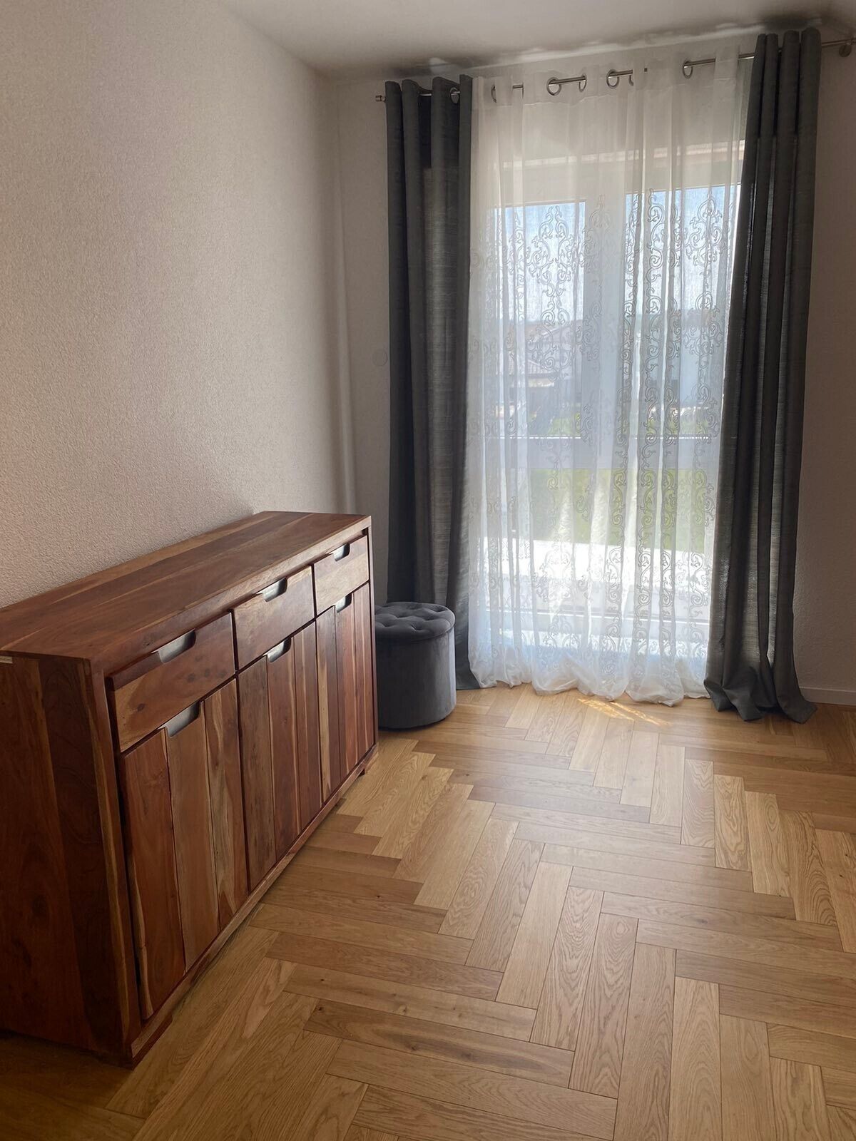 A room with a wooden herringbone floor, a wooden cabinet with drawers, a grey ottoman, and a window with sheer white curtains and dark grey drapes.