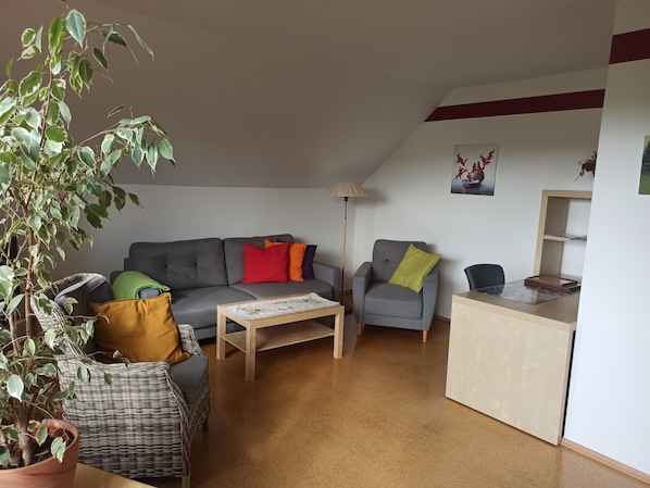 Un salon avec un canapé gris, une petite table, une chaise, une plante en pot et un bureau avec une lampe.