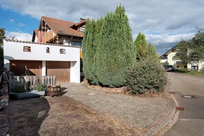 Una calle residencial con pavimento adoquinado, una casa blanca con puerta marrón y un alto árbol de hoja perenne verde.