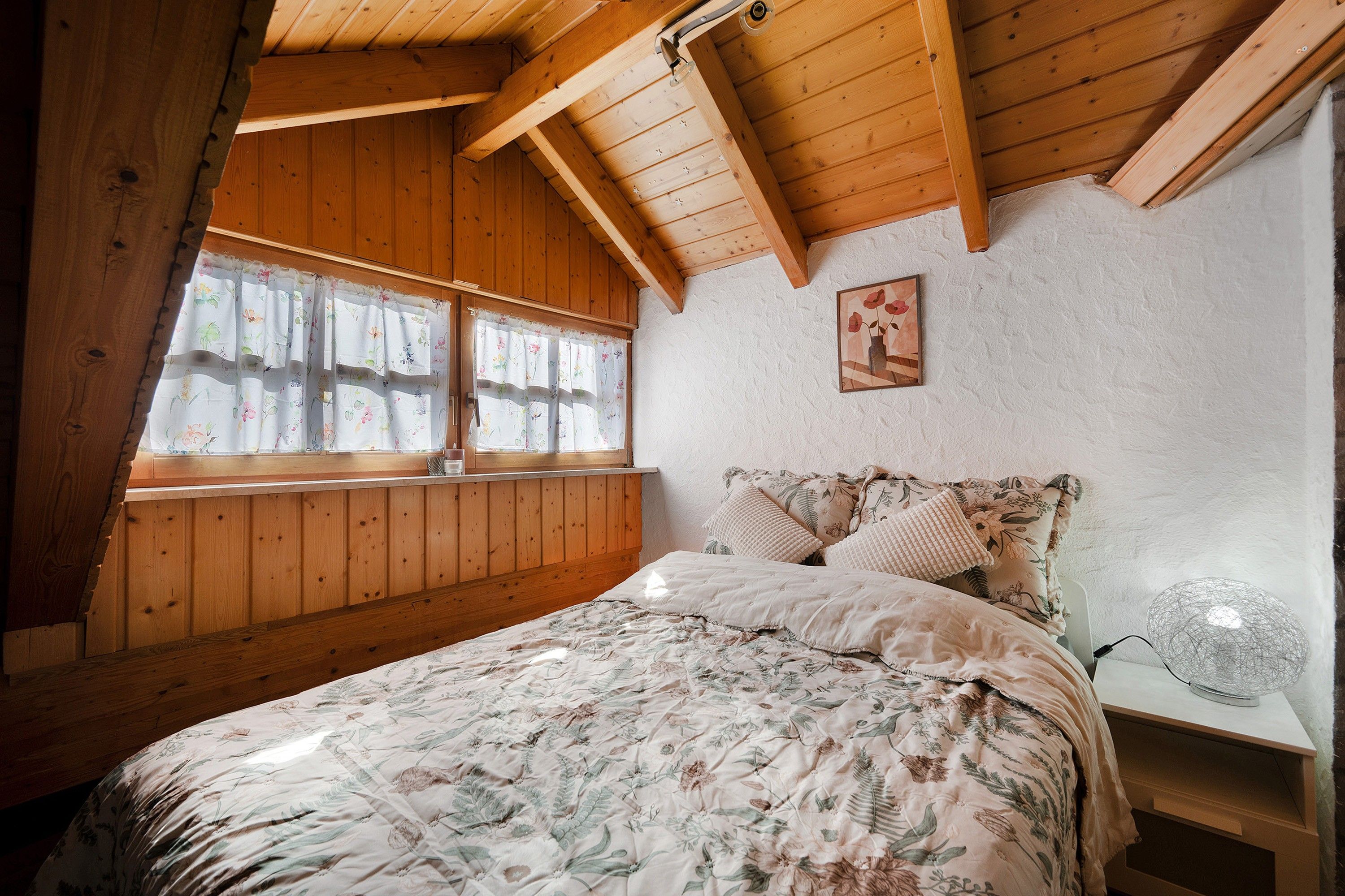 Een slaapkamer met een schuine kap, houten wanden, een bed met bloemenbeddels, een nachtkastje met een lamp en een raam met bloemen gordijnen.