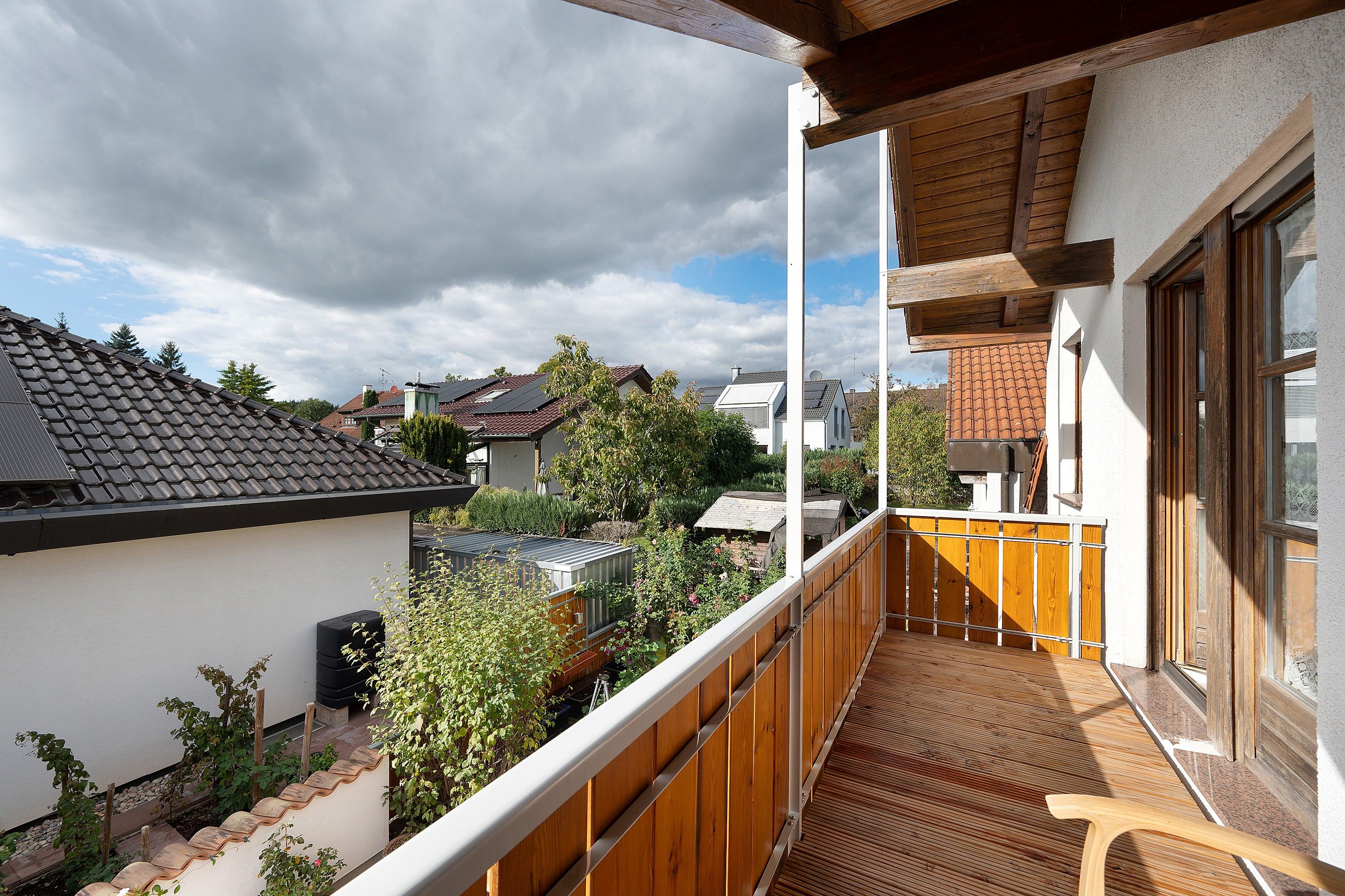 A balcony with a wooden floor and railing, overlooking a residential area with houses and a garden.
