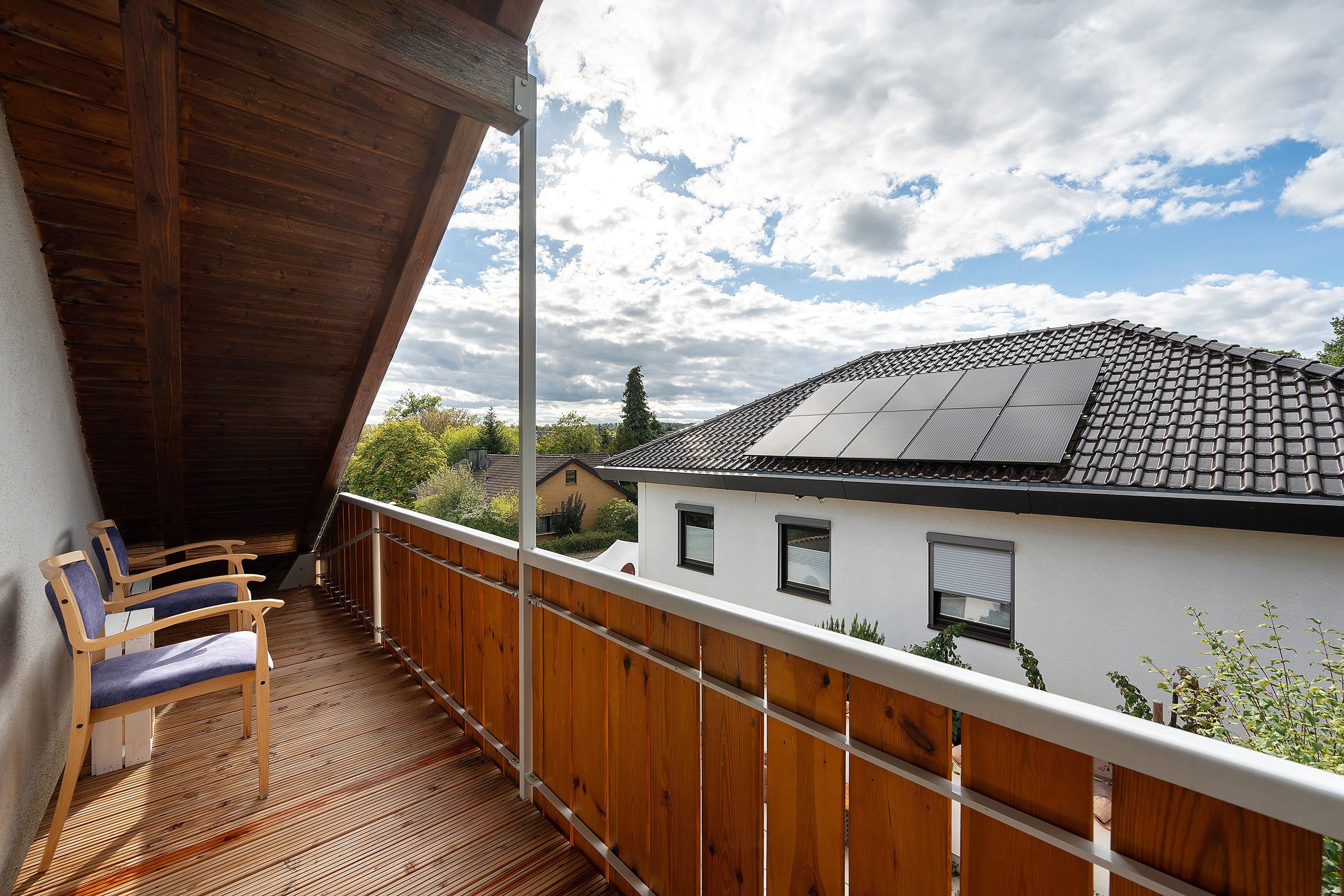 Un balcon en bois avec des chaises, une vue sur une maison équipée de panneaux solaires et un ciel nuageux.