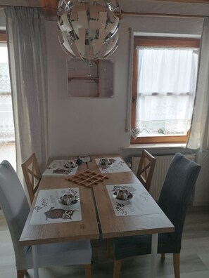 A dining area with a wooden table, four chairs, a chandelier, and a window with curtains.
