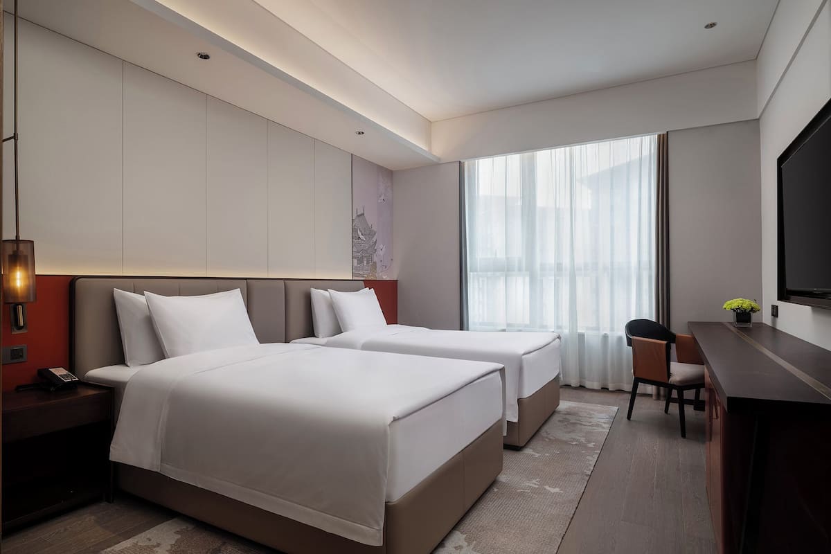 A modern hotel room with two beds, a desk, a chair, and a large window.