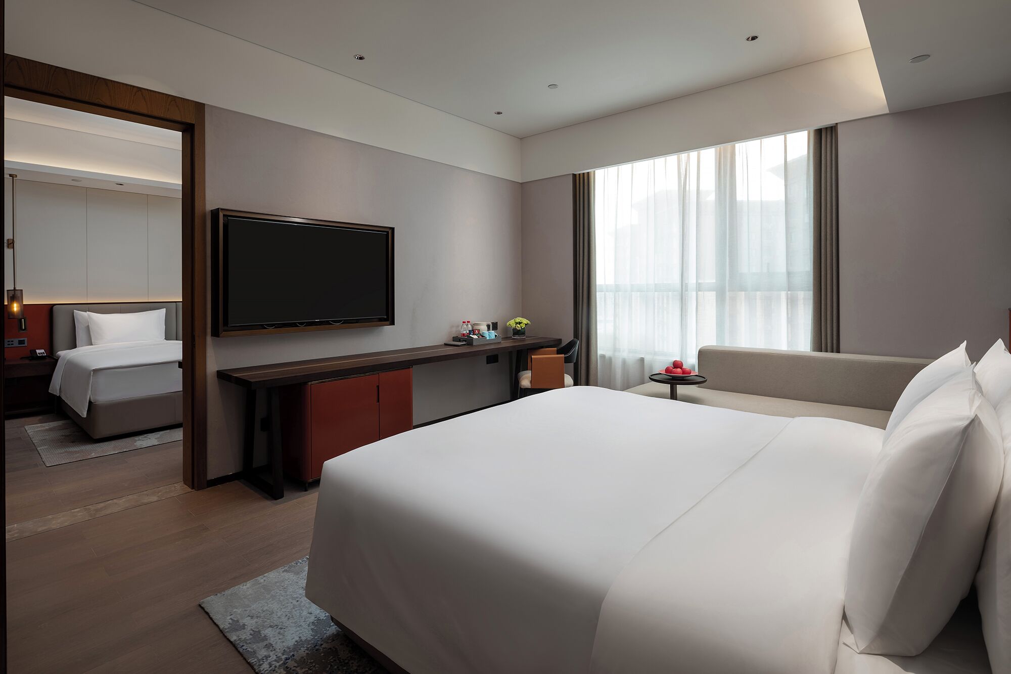 A modern hotel room with a large bed, a flat-screen TV mounted on the wall, a desk with a chair, and a sofa.