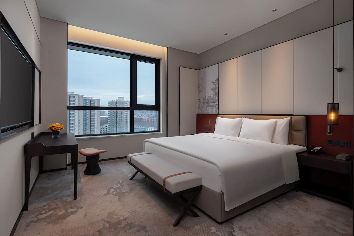 A modern hotel room with a large bed, a desk, a bench, and a view of the city through a large window.