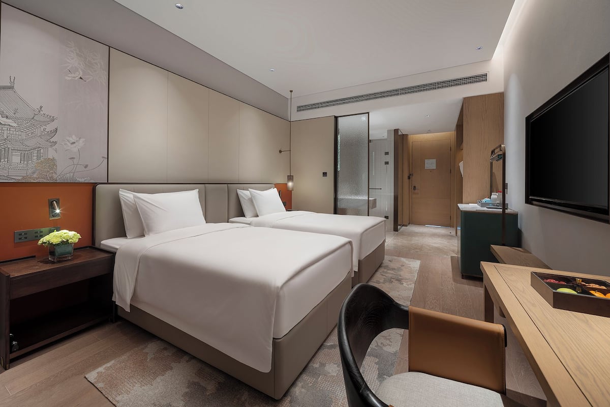 A modern hotel room with two beds, a desk, a TV, and a bathroom visible through a glass door.
