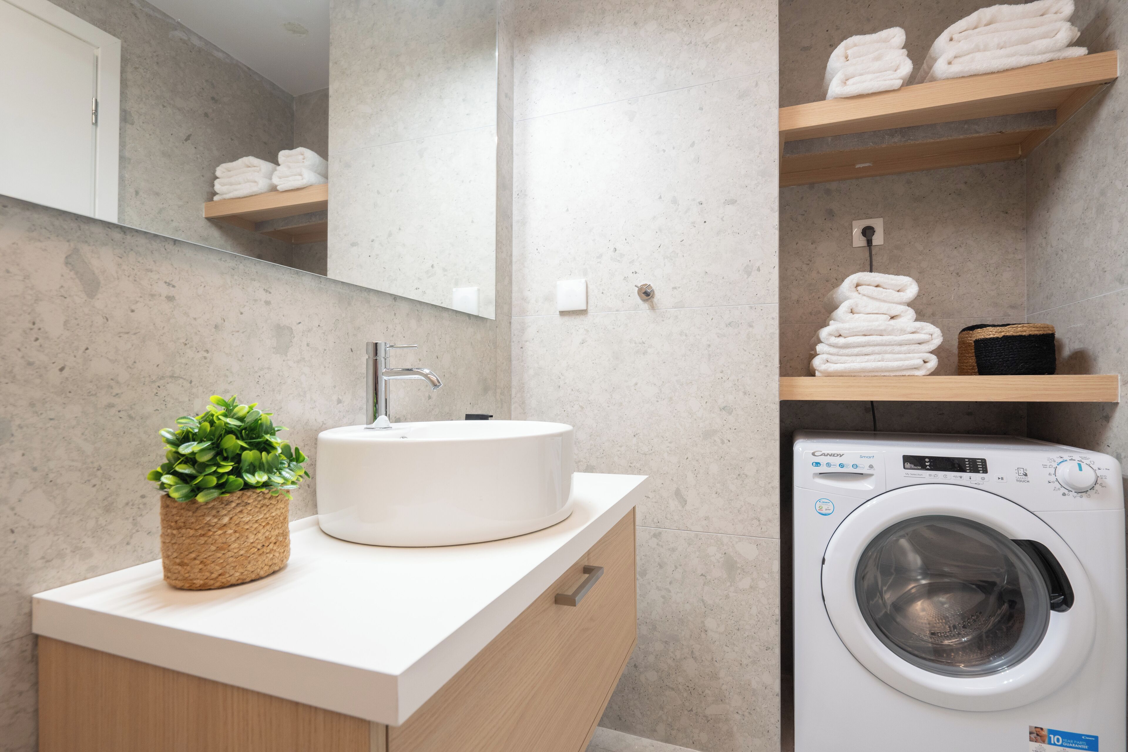 Une salle de bain moderne avec un lavabo rond, un miroir, une petite plante, un lave-linge et des serviettes soigneusement pliées et rangées sur des étagères.