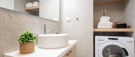 Une salle de bain moderne avec un lavabo rond, un miroir, une petite plante, un lave-linge et des serviettes soigneusement pliées et rangées sur des étagères.