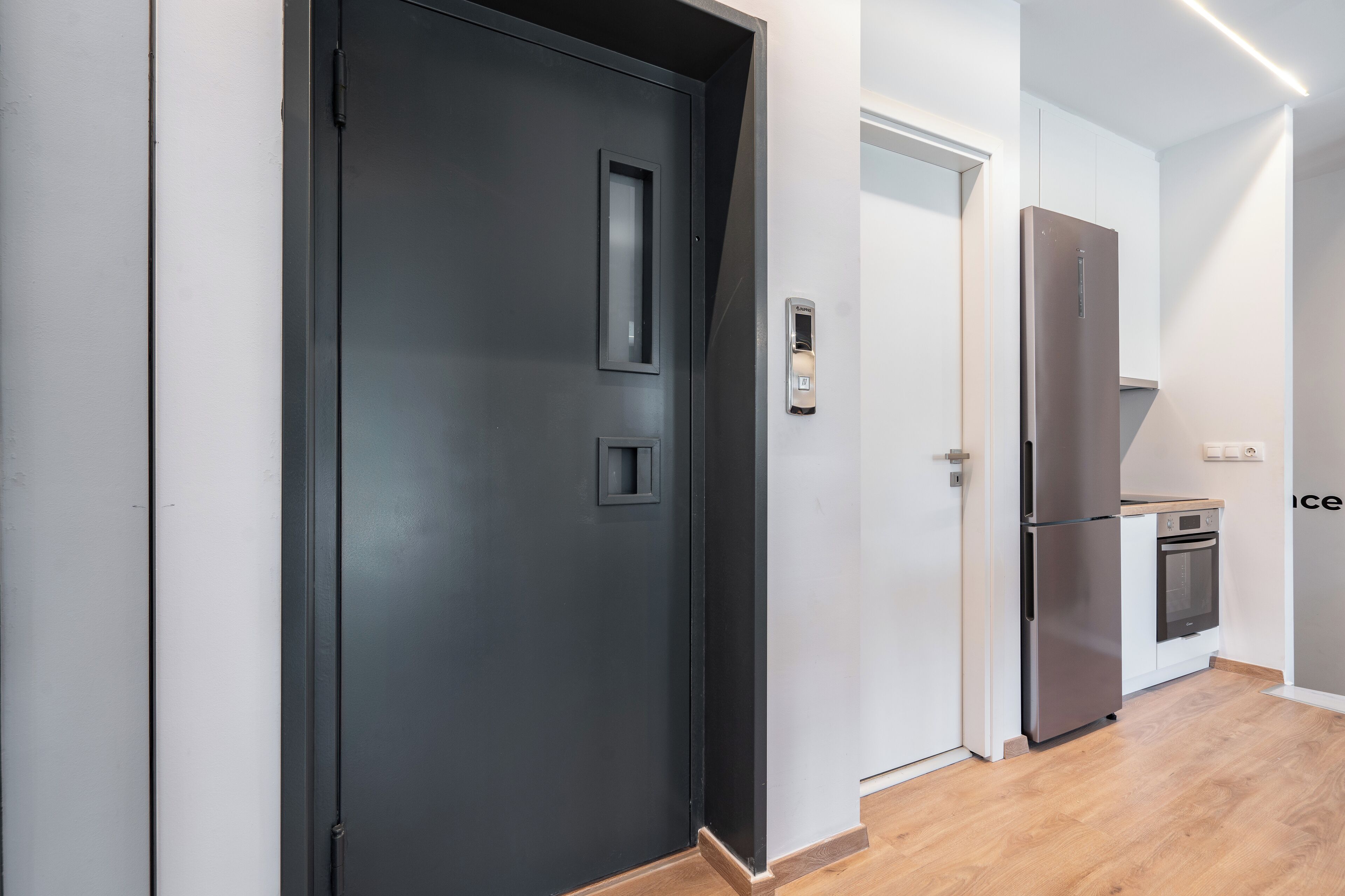 A modern kitchen with a stainless steel refrigerator and oven, a wooden floor, and a dark door with a handle.