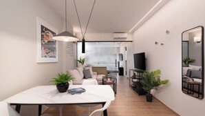 Una sala moderna con un sofá blanco, una mesa de comedor con sillas y un televisor. Hay plantas en el suelo y un espejo grande en la pared.