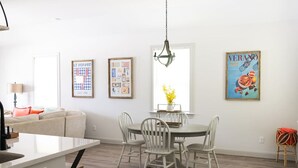 Et moderne køkken med hvide skabe, et spiseområde med et rundt bord og fire stole, samt en stue med sofa og lampe.