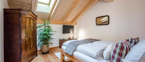 A bedroom with a wooden ceiling, a bed, a wardrobe, a plant, and a TV on the wall.
