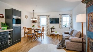 Eine moderne Küche mit dunklen Schränken und einem eingebauten Ofen, ein Essbereich mit Holzsesseln und einem Tisch, ein gemütliches Wohnzimmer mit einer Couch und einer Lampe sowie ein großes Fenster mit Vorhängen.