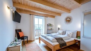 Une chambre avec un lit en bois, un téléviseur fixé au mur, une chaise, une petite table avec une carafe et des verres, et une fenêtre avec des rideaux.