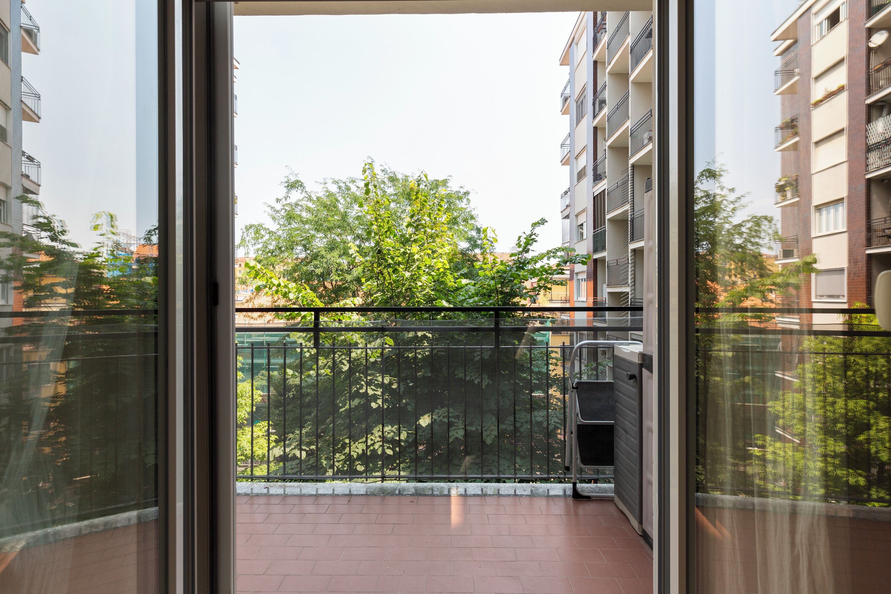 A balcony with a metal railing, a tiled floor, and a view of trees and buildings in the background.